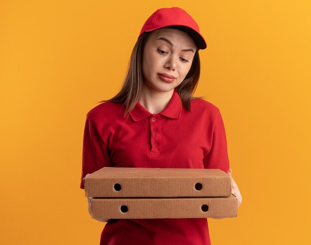 Impressed pretty delivery woman in uniform holds and looks at pizza boxes isolated on orange wall with copy space