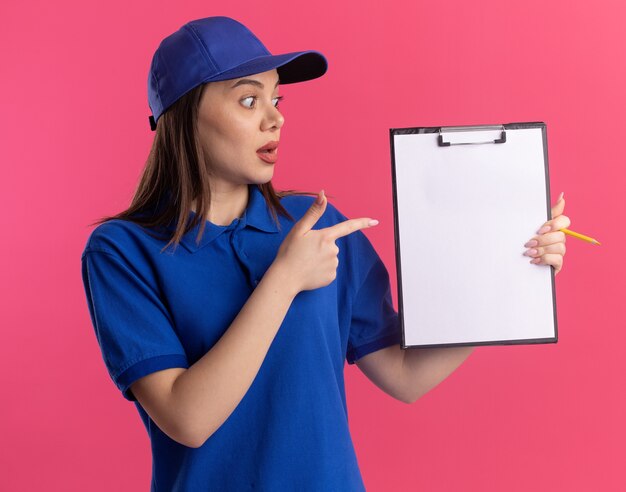 Impressed pretty delivery woman in uniform looks and points at clipboard on pink