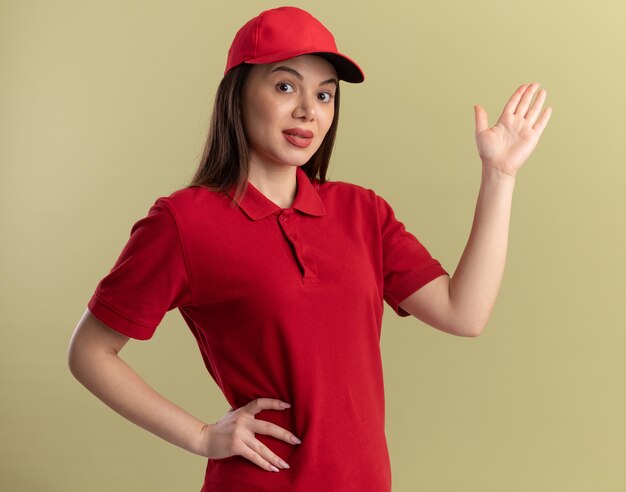 Impressed pretty delivery woman in uniform stands with raised hand on olive green