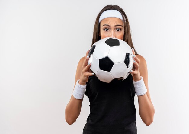 머리띠와 팔찌를 착용하고 축구 공을 들고 복사 공간이 흰 벽에 고립 된 뒤에 숨어있는 인상적인 꽤 스포티 한 소녀