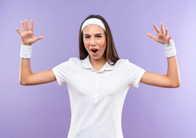 Impressed pretty sporty girl wearing headband and wristband raising hands isolated on purple wall