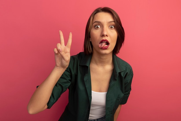 Impressed pretty woman stucks out tongue and gestures victory hand sign isolated on pink wall