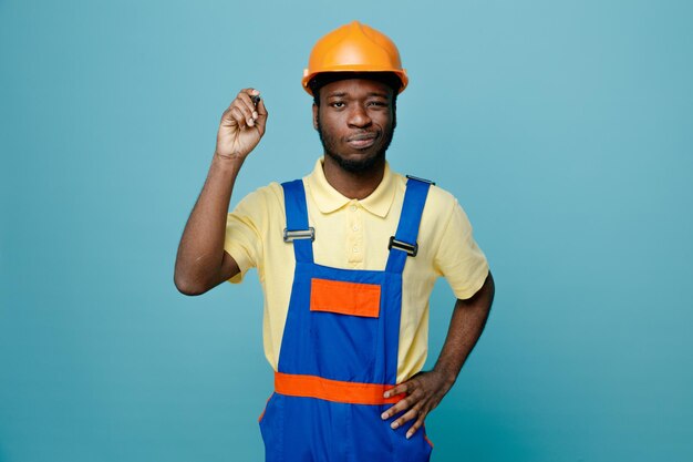 Impressed putting hand on hips young african american builder in uniform holding marker isolated on blue background