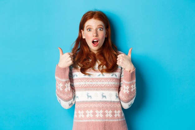 Impressed redhead girl in sweater showing thumbs-up, open mouth fascinated, approve and like product, praising something, standing over blue background.