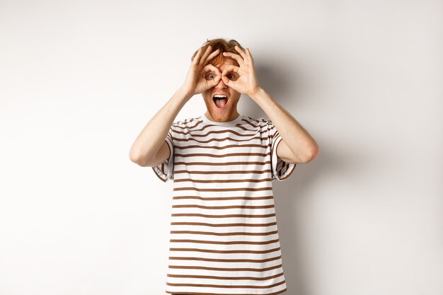 Impressed redhead man checking out promo offer, looking at camera from hand binoculars, smiling amazed, white background