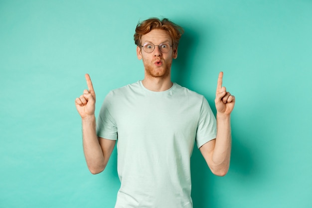 Impressed redhead man in glasses and t-shirt, checking out promo offer, pointing fingers up at copy space, staring at camera amazed, standing over turquoise background