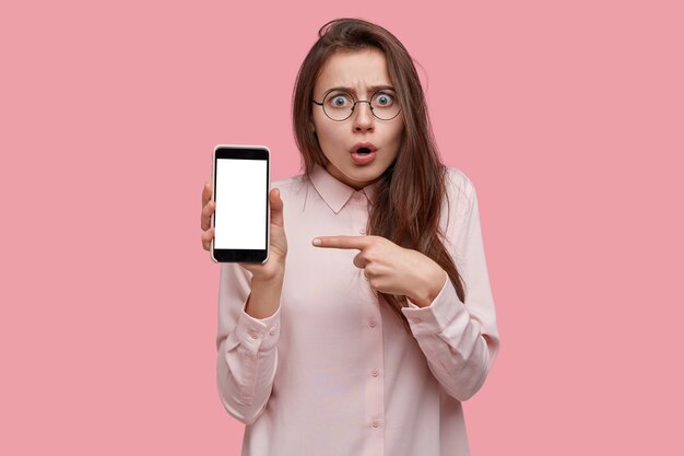 Impressed scared woman opens mouth from interest and thrill, points at mobile phone with empty screen, feels fascinated, dressed in elegant clothes