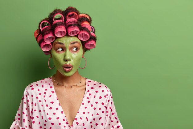 impressed shocked woman applies fresh clay mask for healthy skin, does beauty procedure and facial treatments, wears hair curlers and domestic robe, blank space on green wall.