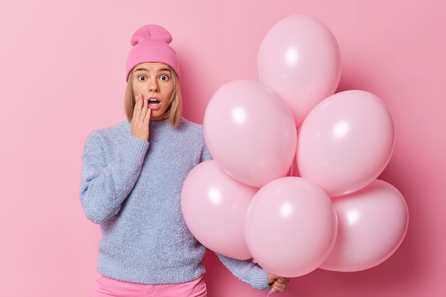 Free photo impressed shocked woman keeps mouth opened looks with disbelief canot believe her eyes wears hat and jumper poses with bunch of inflated balloons isolated over pink background. celebration concept