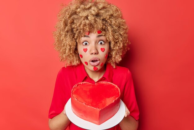 Vaentines Day에 남편을 위해 맛있는 하트 모양의 케이크를 구운 충격을 받은 젊은 여성은 입을 크게 벌리고 카메라를 바라보는 눈으로 빨간색 배경에서 격리된 휴가를 준비합니다