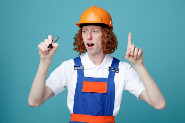 Впечатлен показом номера молодого человека-строителя в униформе, протягивающего маркер к камере на синем фоне
