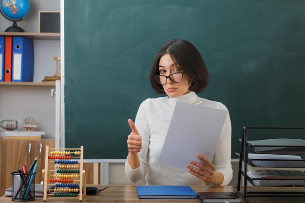 교실에서 학교 도구를 들고 책상에 앉아 종이를 들고 안경을 쓴 젊은 여교사에게 엄지손가락을 치켜드는 인상을 받았다