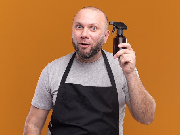 Impressed slavic middle-aged male barber in uniform holding water spray bottle isolated on orange wall