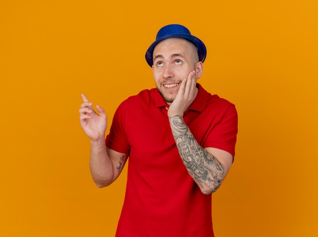 Impressed smiling young handsome party guy wearing party hat keeping hand on face looking and pointing up isolated on orange wall