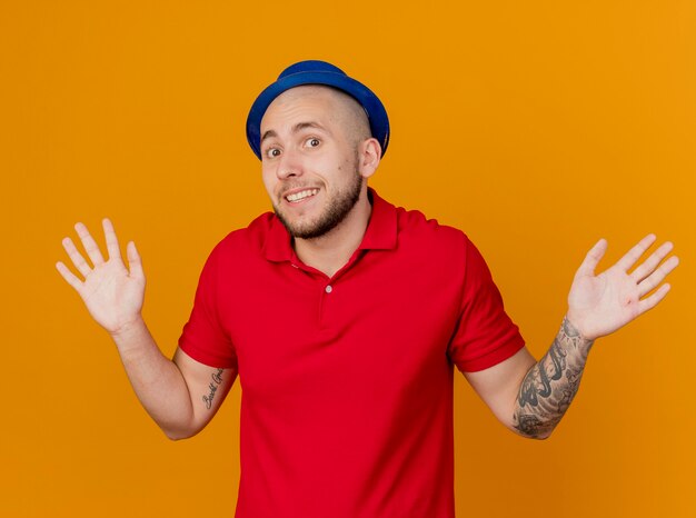 Впечатленный улыбающийся молодой красивый славянский тусовщик в партийной шляпе, смотрящий в камеру, показывая пустые руки, изолированные на оранжевом фоне