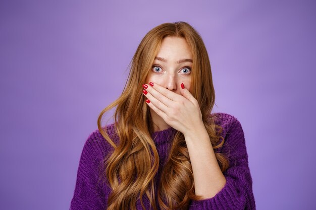 Impressed speechless cute ginger girl hearing stunning gossip covering mouth form amazement and shook raising eyebrows wondered as reacting to unexpected revelation or rumor over purple background.