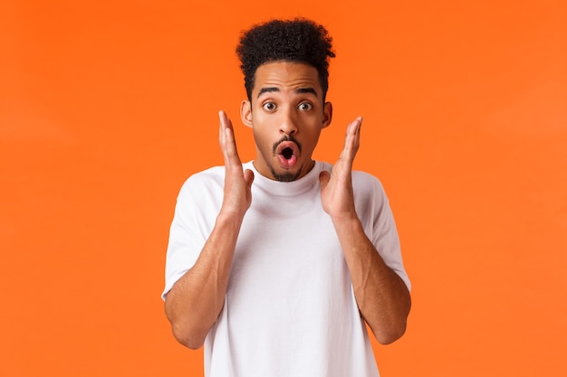 Impressed, startled african-american guy seeing something awesome, gasping wondered and amused, checking-out discounts, season sale, attend stunning performance, standing orange background.