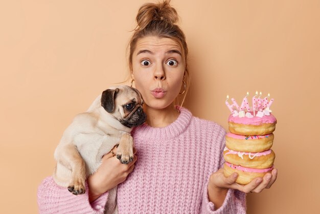 Impressed suprised young woman stares with widely opened eyes holds pug dog on hands poses with festive tasty doughnuts keeps lips folded wears knitted sweater isolated over brown background