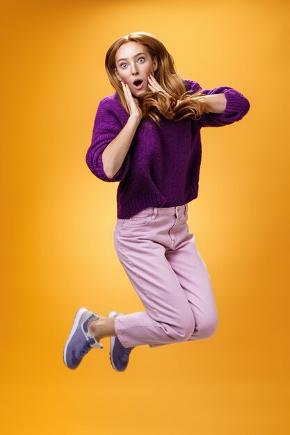 Impressed and surprised amazed good-looking redhead female in purple sweater open mouth astonished and holding hands on cheeks as jumping popping eyes amused over orange background