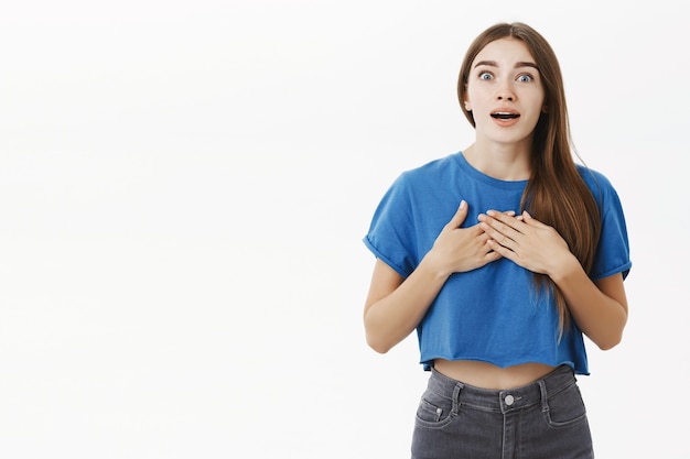 impressed surprised good-looking caucasian female brunette in trendy blue cropped top holding hands on breast opening mouth from excitement and gazing amazed