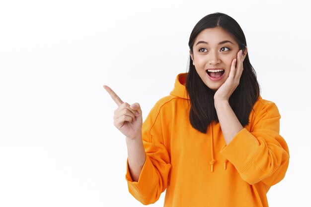 Impressed and surprised kawaii asian girl in orange hoodie touching cheek in awe smiling and pointing left at something amazing cool promo awesome discount offer stand white background