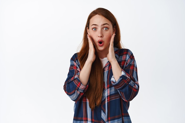 La donna dai capelli rossi impressionata e sorpresa ascolta fantastiche notizie interessanti, dì wow, fissa con soggezione ed eccitazione la fotocamera, tieni le mani sul viso, sfondo bianco