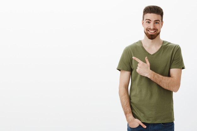 Impressed and surprised very happy bearded guy with deep blue eyes in t-shirt pointing at upper left corner or behind with pleased excited smile telling about incredible product over white wall