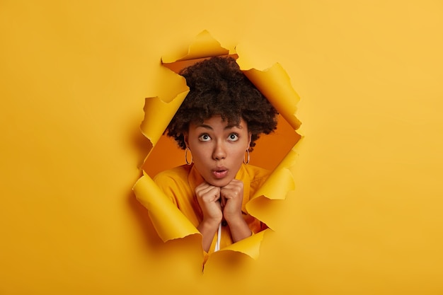Impressed surprised young woman, keeps hands pressed together under chin, poses in torn yellow paper hole background
