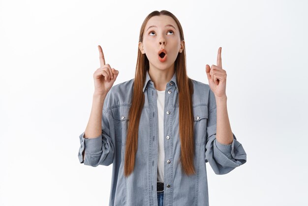Impressed teenage girl female student make wow face staring and pointing up at top advertisement checking out something interesting white background