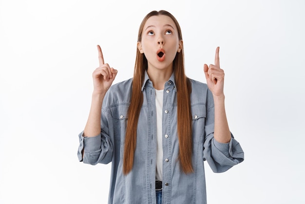 Impressed teenage girl female student make wow face staring and pointing up at top advertisement checking out something interesting white background