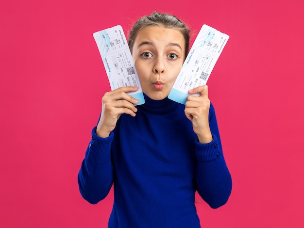 Бесплатное фото Впечатленная девочка-подросток держит билеты на самолет, касаясь ими лица поджатыми губами, изолированными на розовой стене