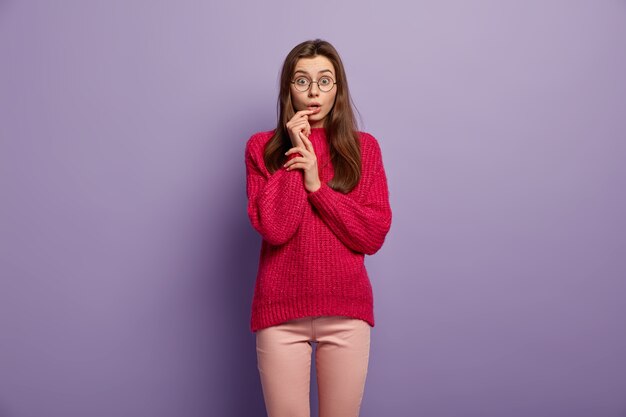 Impressed teenage girl keeps hand near opened mouth, stares with astonishment, wears round spectacles, red ovrsized jumper, isolated over purple wall People, reaction, facial expressions