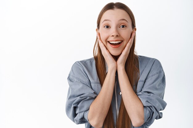 Impressed teenage girl look with fascinated face, admire something beautiful, look at amazing awesome thing, smiling excited, standing over white wall