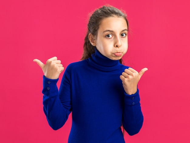 Impressed teenage girl looking at camera puffing cheeks pointing at sides isolated on pink wall