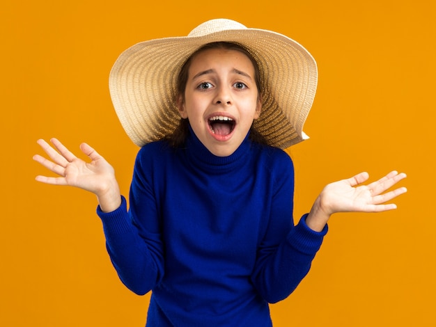 Impressed teenage girl wearing beach hat looking at front showing empty hands isolated on orange wall