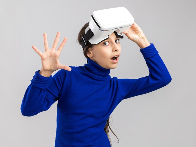 Impressed teenage girl wearing vr headset raising it looking at front showing five with hand isolated on white wall