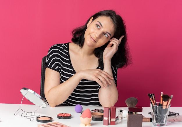 Impressed tilting head young beautiful girl sits at table with makeup tools isolated on pink wall