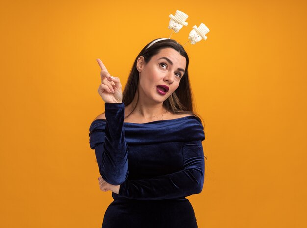 Impressed tilting head young beautiful girl wearing blue dress and christmas hair hoop points at up isolated on orange background