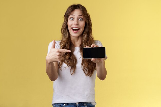 Impressed upbeat happy lucky girl curly long hairstyle open mouth admiration joy like awesome new app show smartphone screen horizontal phone display stand yellow background amazed