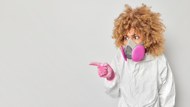 Impressed woman dressed in protective clothes wears respirator points away on copy space checks level of radioactive radiation in danger zone isolated over white background World disaster concept