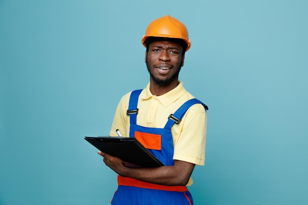 Impressed young african american builder in uniform holding clipboard isolated on blue background
