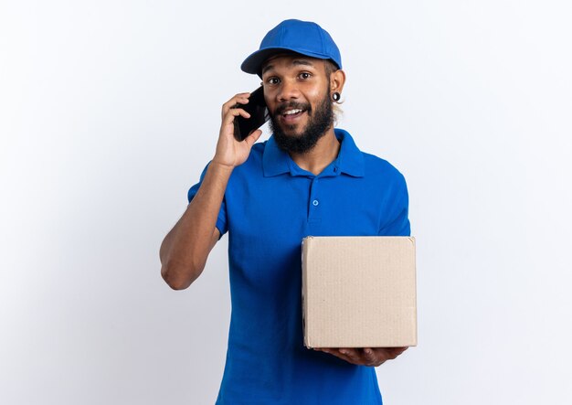 Impressed young afro-american delivery man holding cardboard box talking on phone isolated on white background with copy space