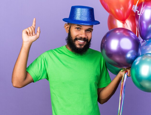 Impressed young afro-american guy wearing party hat holding balloons points at up 