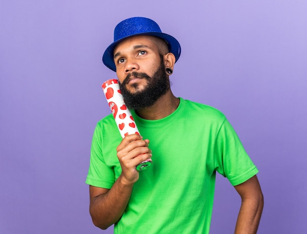 Впечатленный молодой афро-американский парень в партийной шляпе, держащий пушку с конфетти, изолированную на синей стене