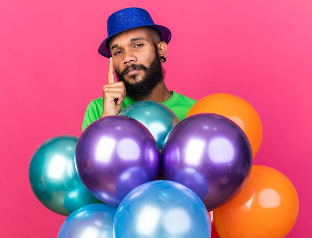 Impressed young afro-american guy wearing party hat standing behind balloons 