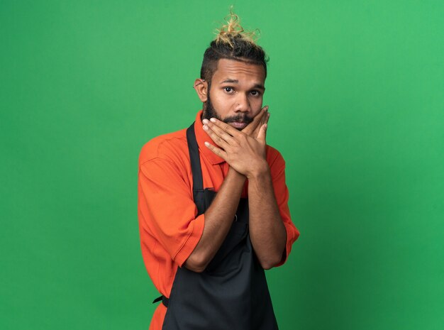 Impressed young afro-american male barber wearing uniform keeping hands crossed on chin 