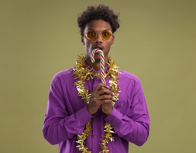 Free photo impressed young afro-american man wearing glasses with tinsel garland around neck holding christmas candy cane near mouth looking at camera isolated on olive green background