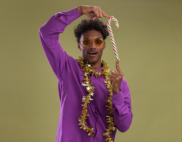 Free photo impressed young afro-american man wearing glasses with tinsel garland around neck holding christmas candy cane vertically looking at camera isolated on olive green background with copy space