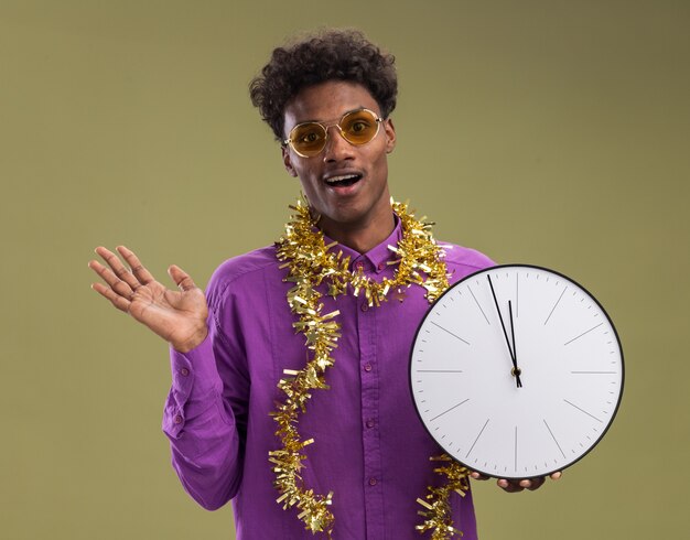 Впечатленный молодой афро-американский мужчина в очках с гирляндой из мишуры на шее держит часы, глядя в камеру, показывая пустую руку, изолированную на оливково-зеленом фоне