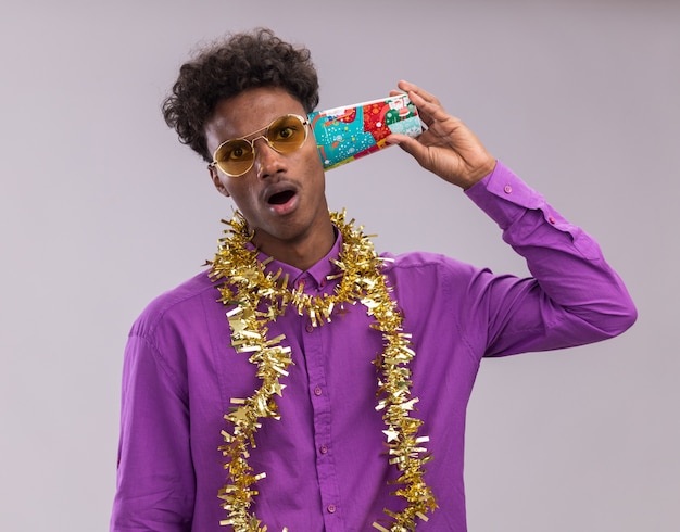 Impressed young afro-american man wearing glasses with tinsel garland around neck holding plastic christmas cup next to ear listening to conversation looking at camera isolated on white background